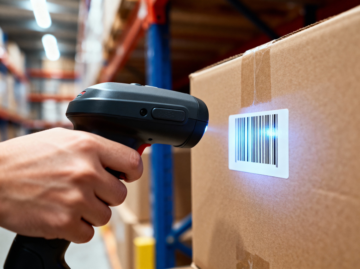Close-up of a worker scanning a barcode on a cardboard box using a handheld scanner in a brightly lit warehouse.